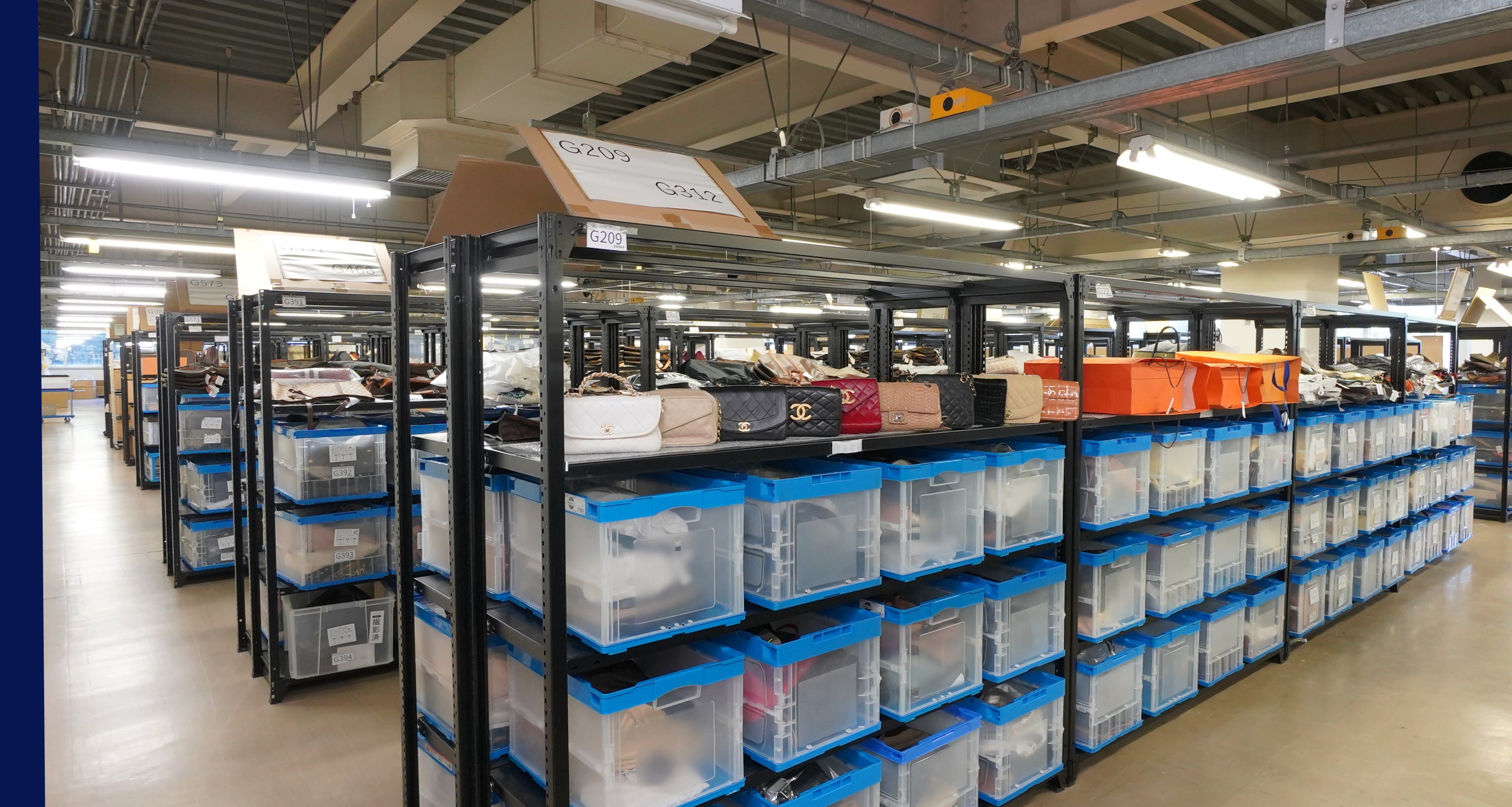 A warehouse storage aisle with tall black shelving units holding many clear plastic bins with blue lids and assorted boxes on top.